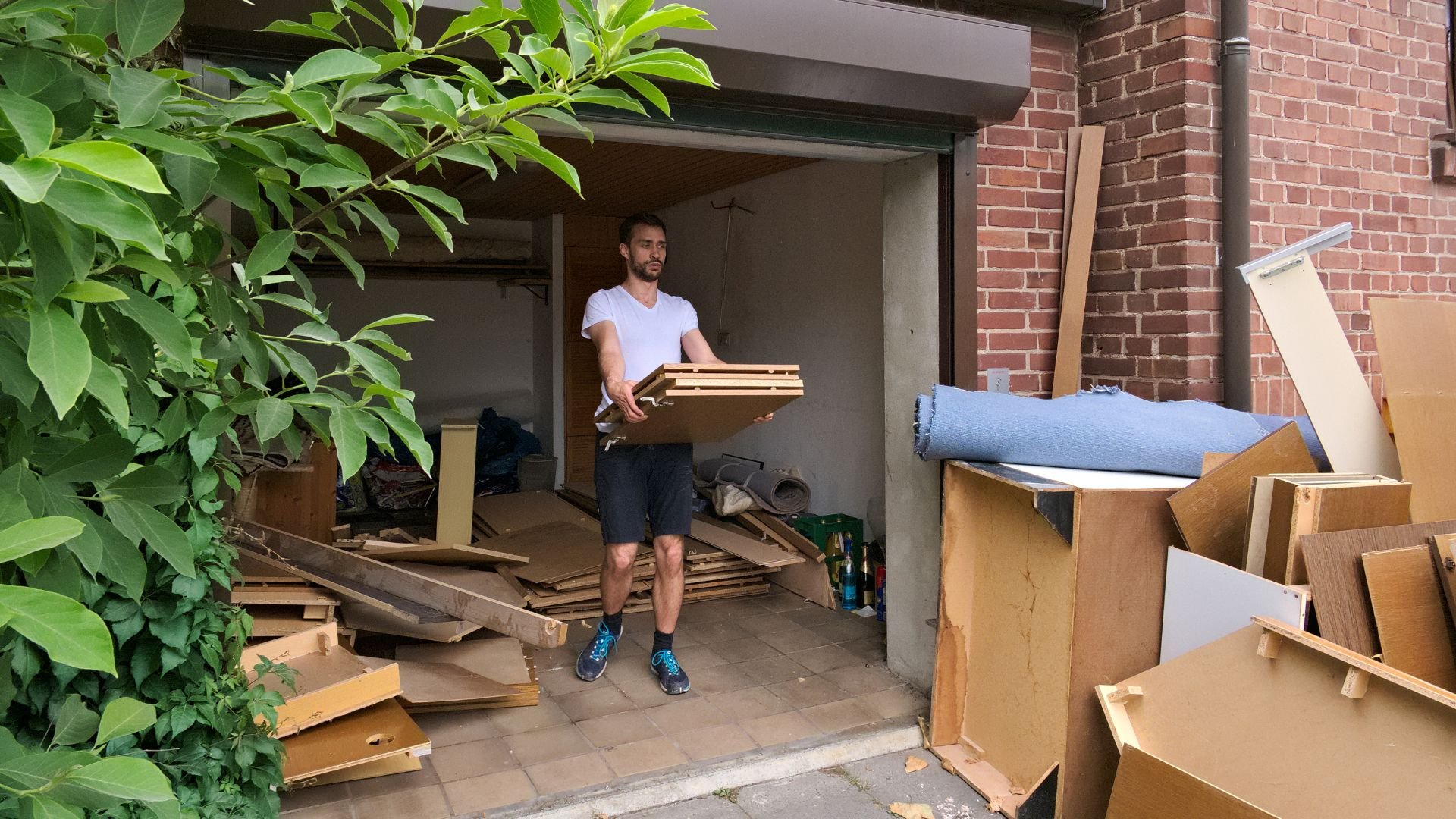 Person moving wooden boards in cluttered storage area with green plants