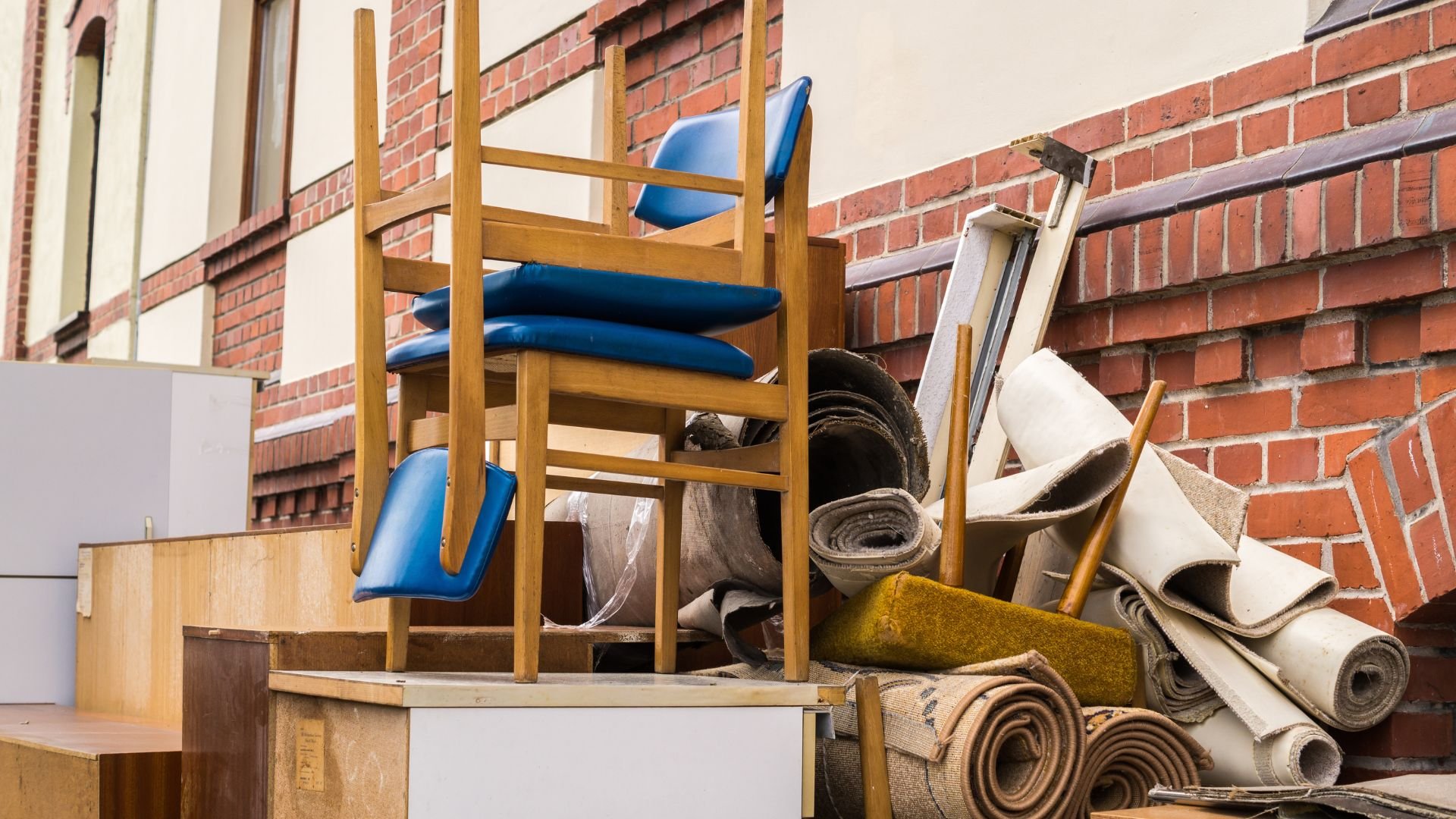 Stacked wooden chairs, carpet rolls, and moving boxes near brick wall