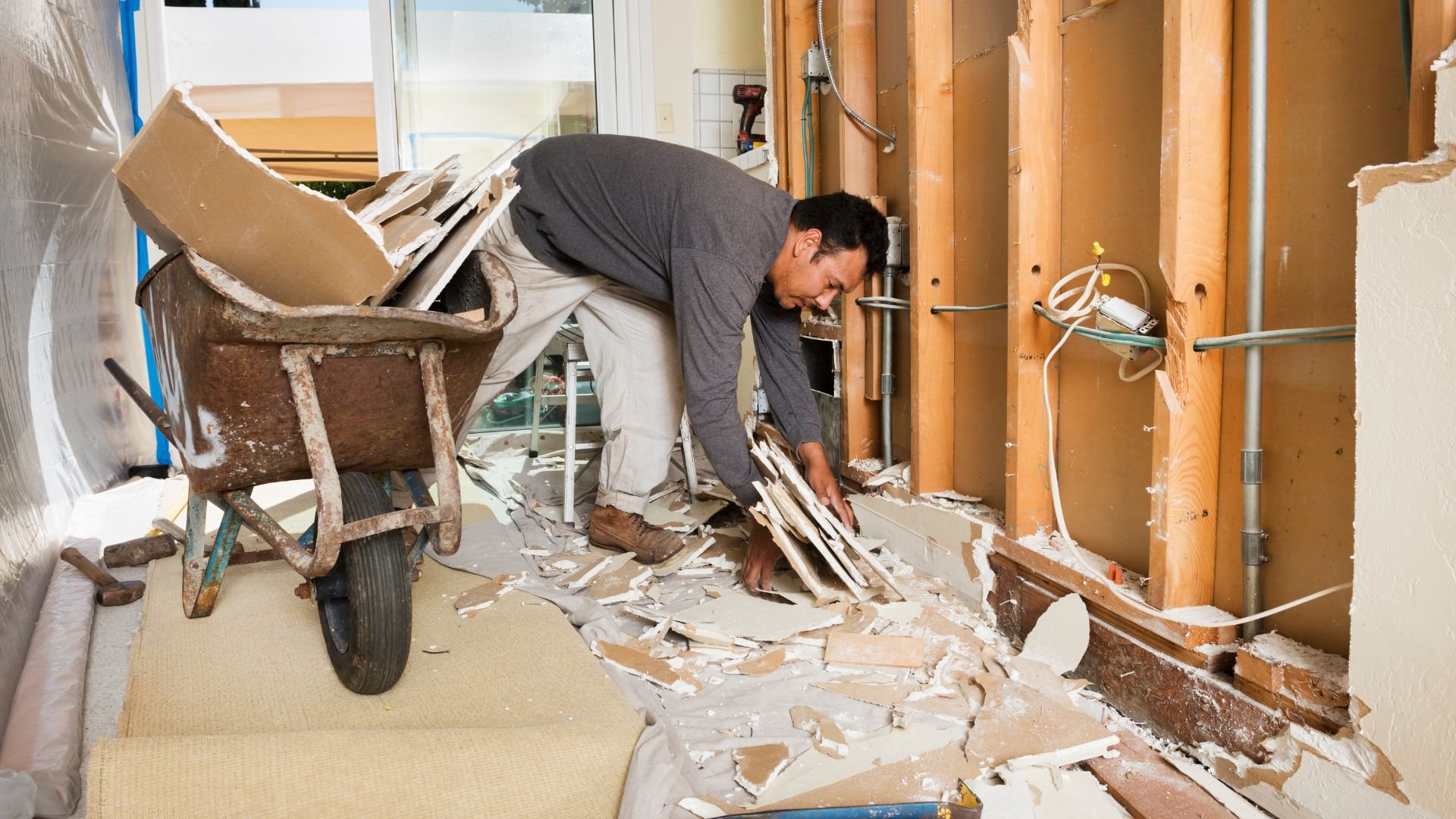 Worker removing drywall during home renovation with wheelbarrow nearby