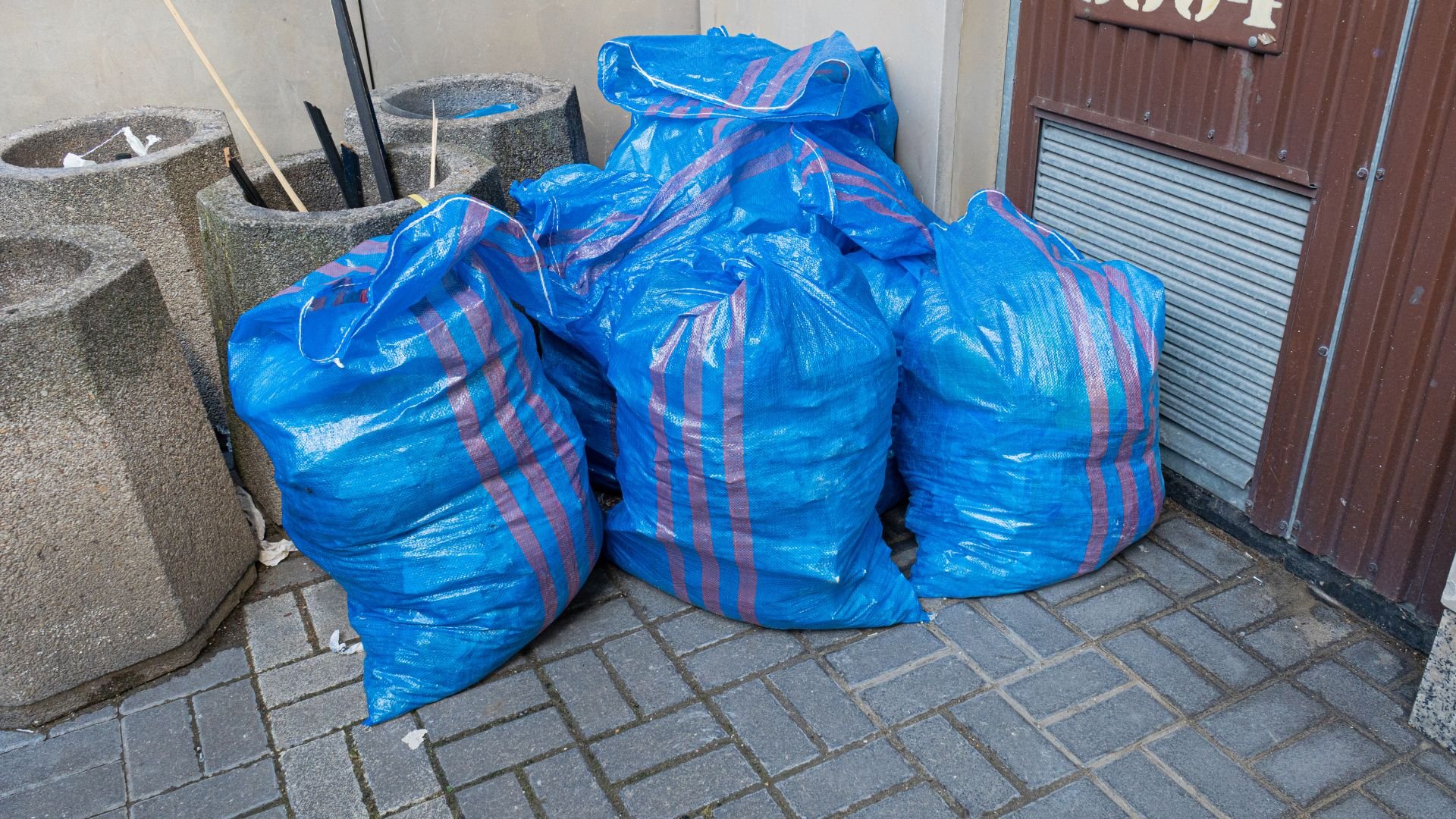 Blue striped garbage bags stacked near concrete planters and a metal door