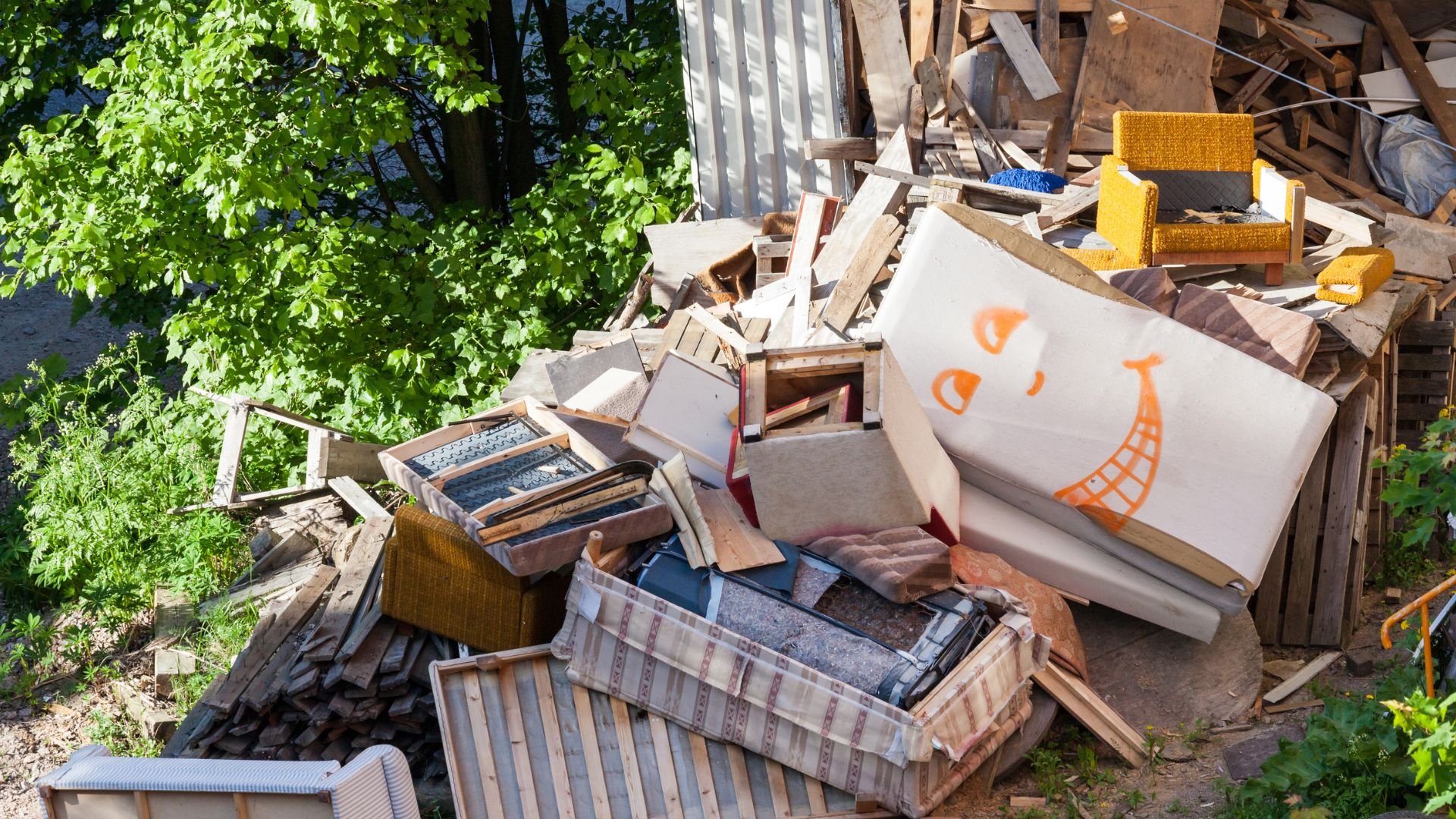 Cluttered pile of wooden debris, furniture, and boxes near green foliage